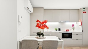 Eine moderne Küche mit weißen Schränken, einer Mikrowelle und einer Kaffeemaschine. Ein Esstisch mit zwei Stühlen und eine Vase mit roten Blumen sind ebenfalls vorhanden. Ein Notausgangsschild ist an der Wand sichtbar.