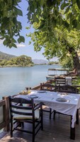 Un área de comedor junto al lago con mesas dispuestas para dos, con vistas a un lago tranquilo y montañas distantes.