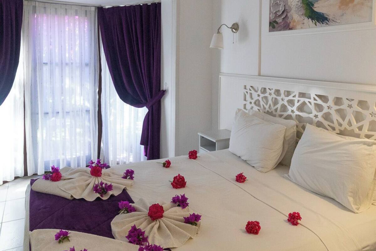 A hotel room with a large bed, a window with curtains, an air conditioning unit, a wall-mounted lamp, and a painting.