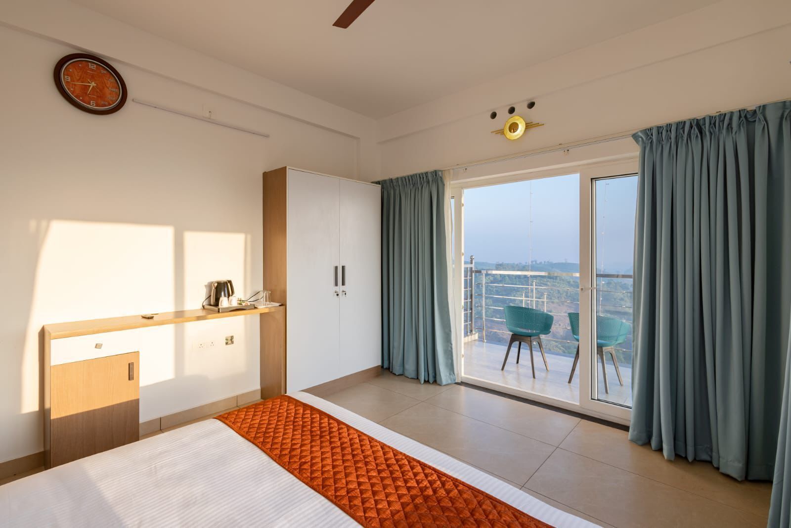 Un dormitorio moderno con cama, armario, escritorio con silla, ventana grande con cortinas y vistas a la ciudad.