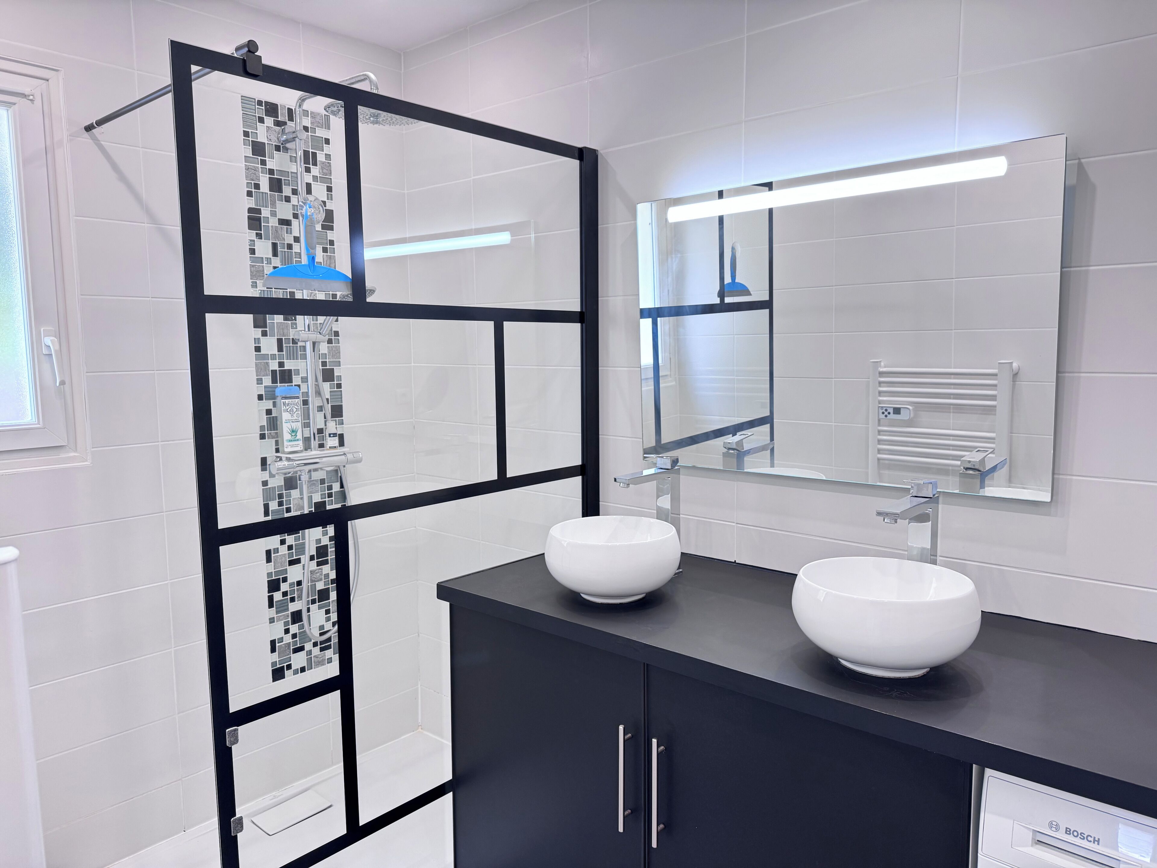 Un baño moderno con dos lavamanos blancos, un espejo grande y una cabina de ducha con marco negro.