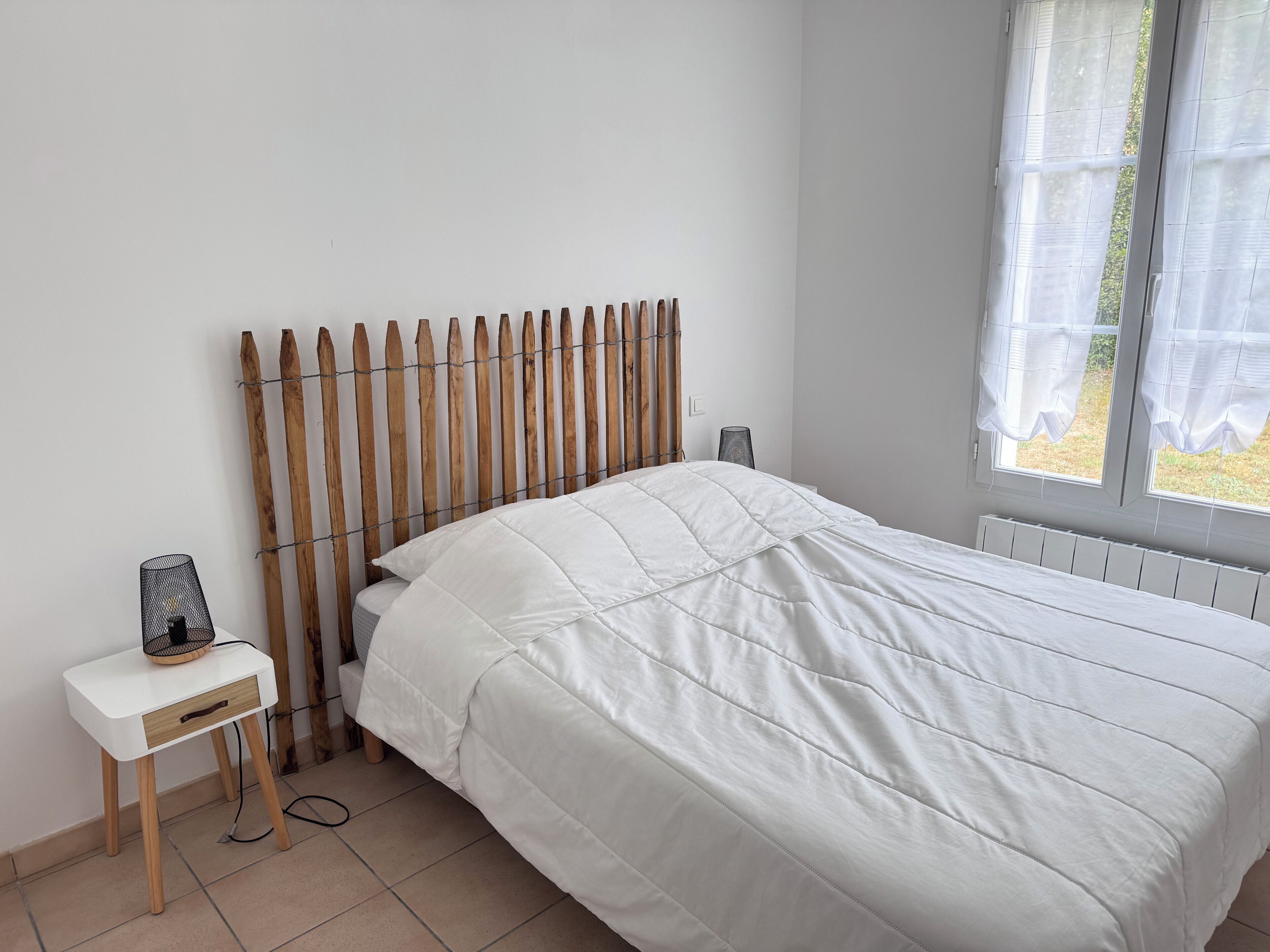 Ein Schlafzimmer mit hölzernem Kopfende, weißer Bettdecke, Nachttisch mit Lampe und einem Fenster mit Vorhängen.
