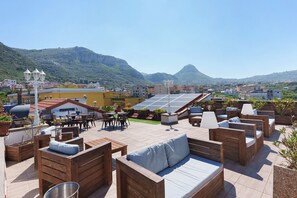 A rooftop patio with wooden furniture, a dining area, and a view of the city and mountains. - Reginella Garden View Studio with Shared Terrace, Wi-Fi, and Air Conditioning (Meta)