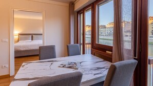 Ein Hotelzimmer mit Bett, Esstisch mit Stühlen, Blick auf einen Fluss und Gebäude sowie einer Tür zu einem weiteren Raum.