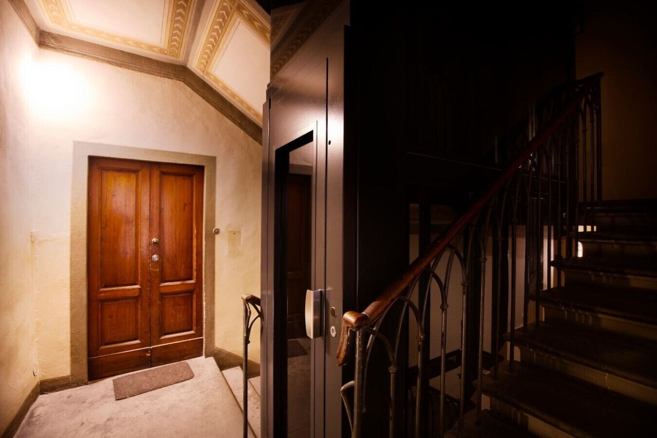 A hallway with a wooden door, a glass elevator, and a staircase with metal railings.