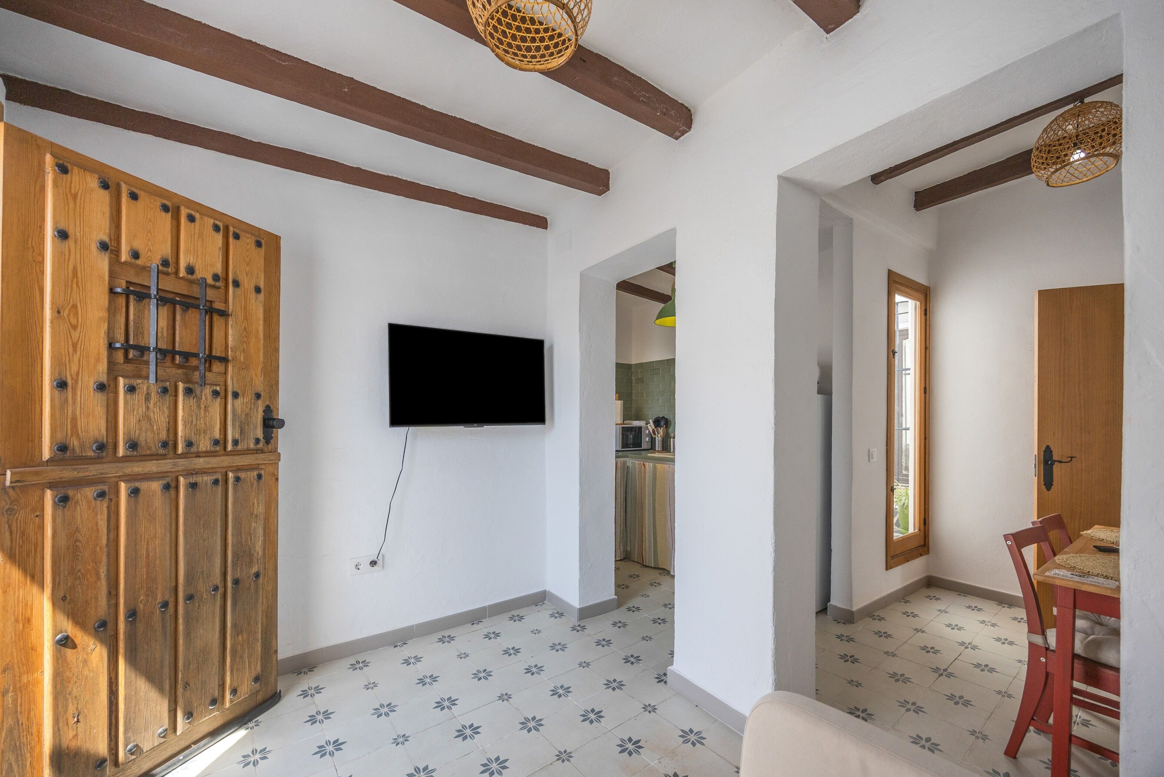 A room with a wooden door, a mounted TV, a dining table with chairs, and a kitchen in the background.