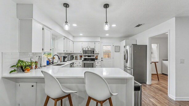 Una cocina moderna con armarios blancos, una isla central con dos taburetes altos, electrodomésticos de acero inoxidable e iluminación colgante.