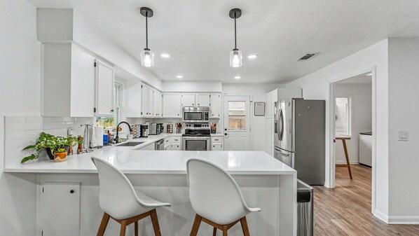 A modern kitchen with white cabinetry, a central island with two bar stools, stainless steel appliances, and pendant lighting. - Conveniently Located - S Lakeland Pool Home, Pet and Family Friendly (Lakeland)