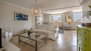 Een woonkamer met een witte bank, een glazen salontafel, een schilderij aan de muur en een groene dressoir.