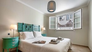 Een keurig opgemaakt bed met een tijdschrift, twee bedlampen, een blauwgroene nachtkastje en een raam met uitzicht op een gebouw.