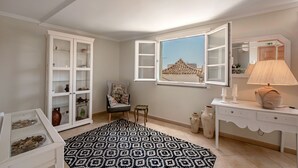 Een kamer met een groot raam, een witte kast met glazen deuren, een kamerbreed tapijt met patroon, een stoel, een bijzettafeltje en een bureau met een lamp.