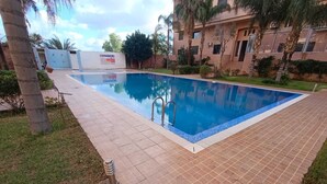 A swimming pool with a tiled deck, surrounded by greenery and a building with a sign. - Beautiful Apartment in Marrakech, GUELIZ (Marrakech)