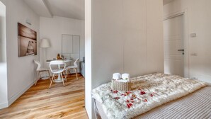 Una stanza con pavimento in legno, pareti bianche, un letto con una coperta bianca e morbida, un tavolino con sedie e una lampada.
