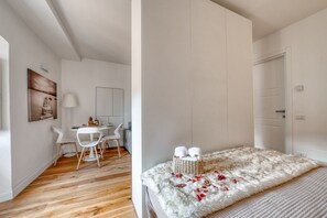 Ein Zimmer mit Holzboden, weißen Wänden, einem Bett mit weißer, flauschiger Decke, einem kleinen Tisch mit Stühlen und einer Lampe.