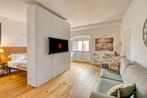 Ein modernes Wohnzimmer mit einem Flachbildfernseher, einem grauen Sofa, Holzboden und einem Essbereich im Hintergrund.