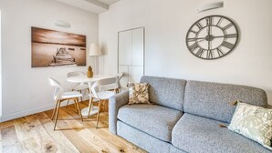 Un soggiorno moderno con un divano grigio, un orologio da parete rotondo, un pavimento in legno e un'area pranzo con sedie bianche e un tavolo.