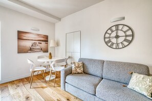 Ein modernes Wohnzimmer mit grauem Sofa, einer runden Wanduhr, Holzboden und einem Essbereich mit weißen Stühlen und einem Tisch.