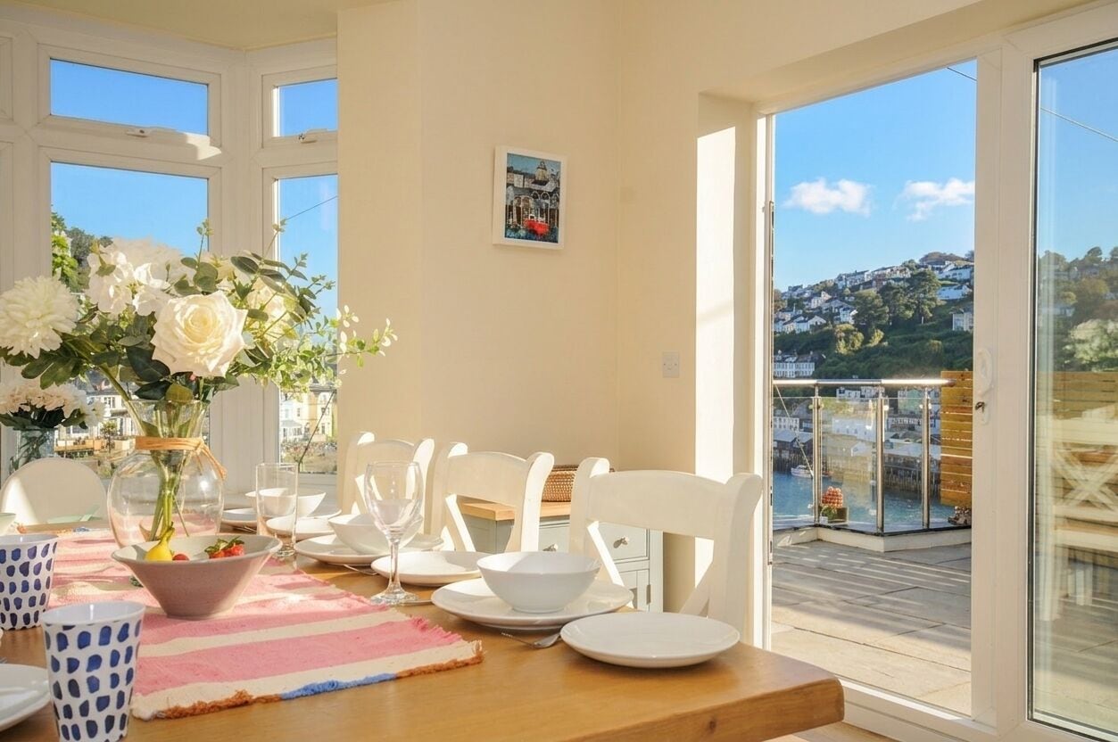 Comedor con una mesa dispuesta para cuatro personas, un jarrón de flores y vistas a un pueblo costero a través de amplios ventanales.