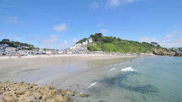 A coastal beach with clear waters, a rocky foreground, and buildings on a hillside in the background.