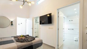 Quarto moderno com cama, televisão, espelho e banheiro visível através de uma porta aberta.