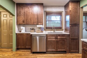A kitchen with wooden cabinets, a stainless steel dishwasher, and a window with blinds.