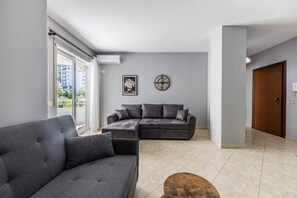 Un salon moderne avec un canapé gris, un canapé plus petit, une table basse en bois et une grande fenêtre donnant sur des immeubles.