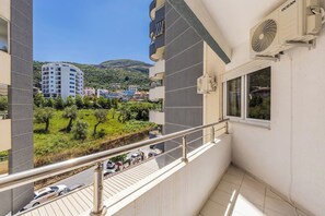 Een balkon met een metalen hekwerk, een airconditioning en uitzicht op een groen landschap met bomen en een heuvel op de achtergrond.