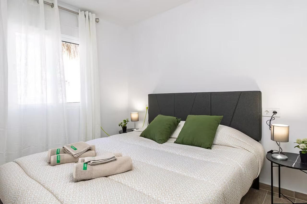 Una cama bien hecha con almohadas, una mesita de noche con lámpara y una ventana con cortinas.
