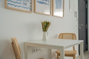 Uma mesa branca com um vaso de plantas secas, duas cadeiras de vime e quadros na parede retratando peixes e ondas.