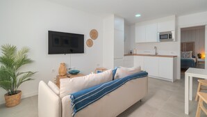 Un salon moderne comprenant un canapé, un téléviseur à écran plat, un coin cuisine avec des placards et un four à micro-ondes, ainsi qu’une table à manger avec des chaises.