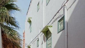 Ein schmaler Gang mit einem weißen Gebäude, das grüne Fensterläden und eine Palme links zeigt.