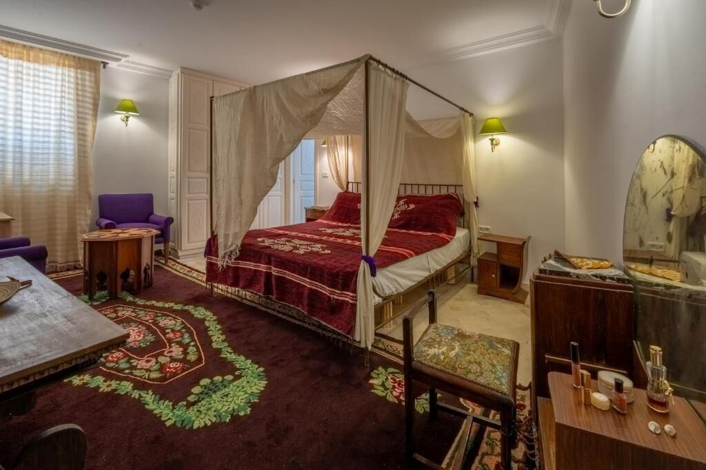 A four-poster bed with a red bedspread, a purple armchair, a wooden side table, and a mirror on the wall.