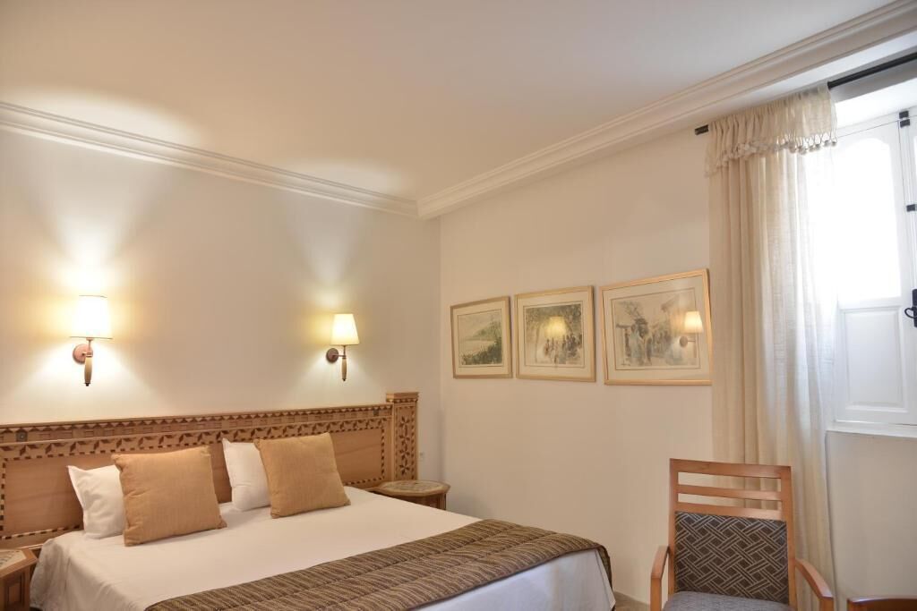 Una camera d'albergo con un letto, due lampade a parete, una sedia e tre quadri appesi alla parete.
