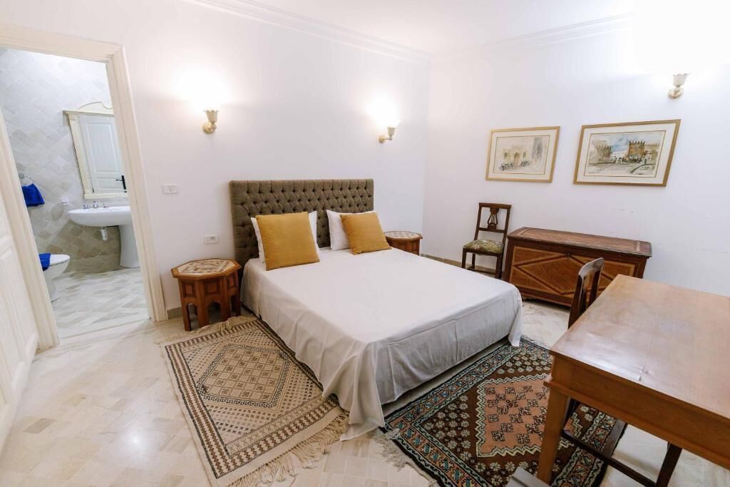 Un dormitorio con una cama, mesitas de noche, un aparador de madera, una pequeña mesa redonda y dos luces fijadas a la pared.
