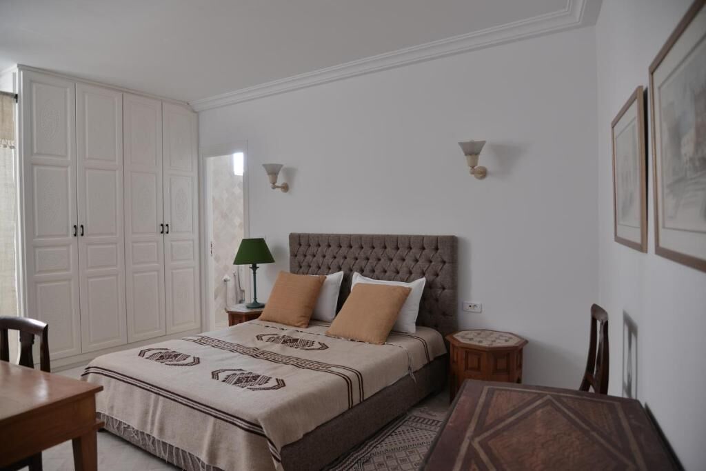 A bedroom with a bed, a nightstand, a lamp, a chair, a table, and a wardrobe.