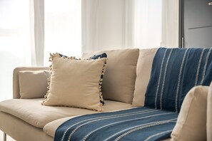 Een beige bank met een blauw-wit gestreepte deken, een beige kussen met franjes, en een effen beige kussen.