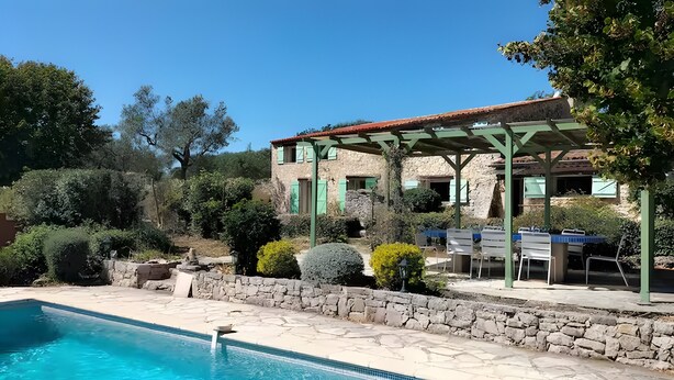 A pool with a stone border, a stone wall, and a building with a green roof and shutters.