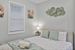 Un dormitorio con cama, mesita de noche, lámpara, ventana con persianas y cuadros en la pared.
