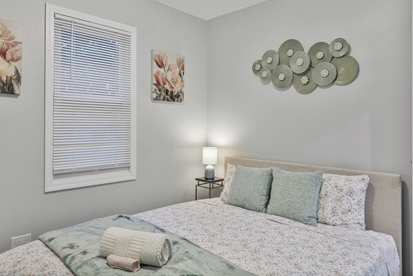 Une chambre à coucher comprenant un lit, une table de chevet, une lampe, une fenêtre avec des stores et des œuvres d’art murales.