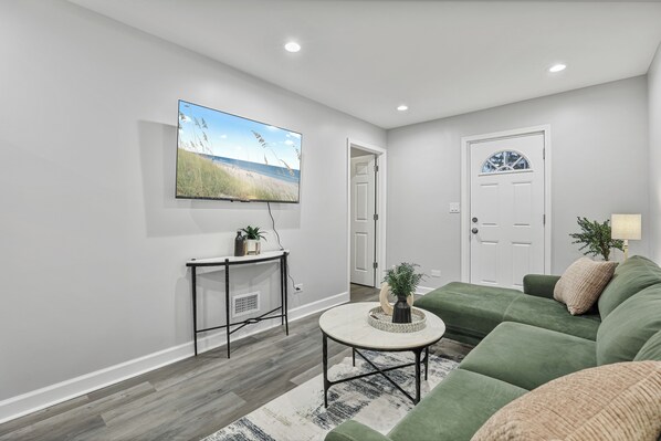 A modern living room with a flat-screen TV, a green sofa, a small coffee table, and a door leading to another room.