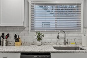 Une cuisine dotée d’un évier en acier inoxydable, d’un plan de travail carrelé blanc et d’une fenêtre avec des stores.