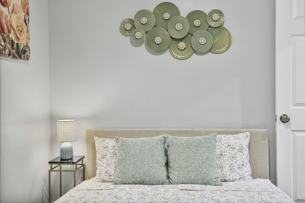 Un dormitorio con cama, almohadas, mesita de noche, lámpara y decoración en la pared.