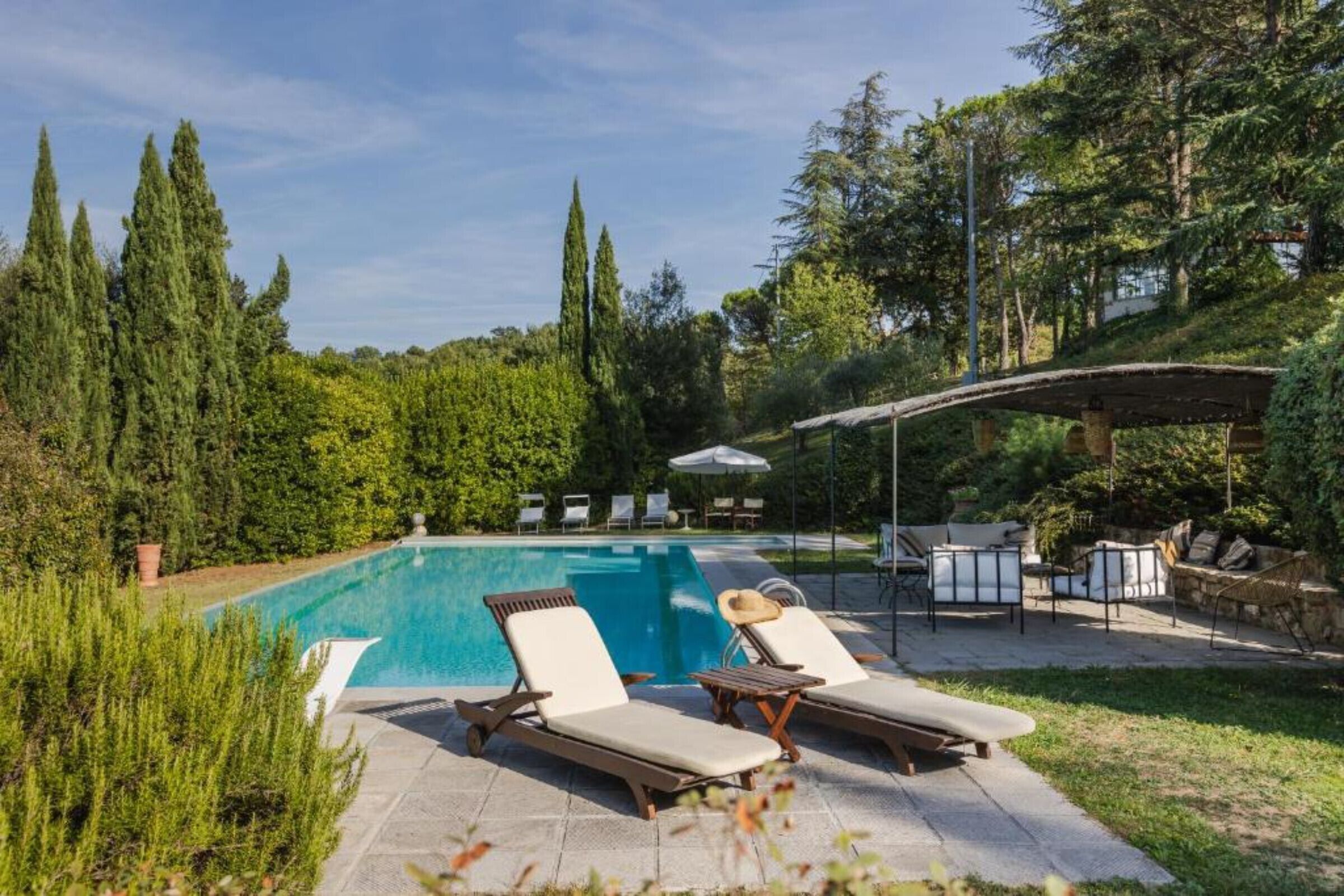 Ein Pool mit Liegestühlen und einer überdachten Sitzgelegenheit, umgeben von Bäumen.