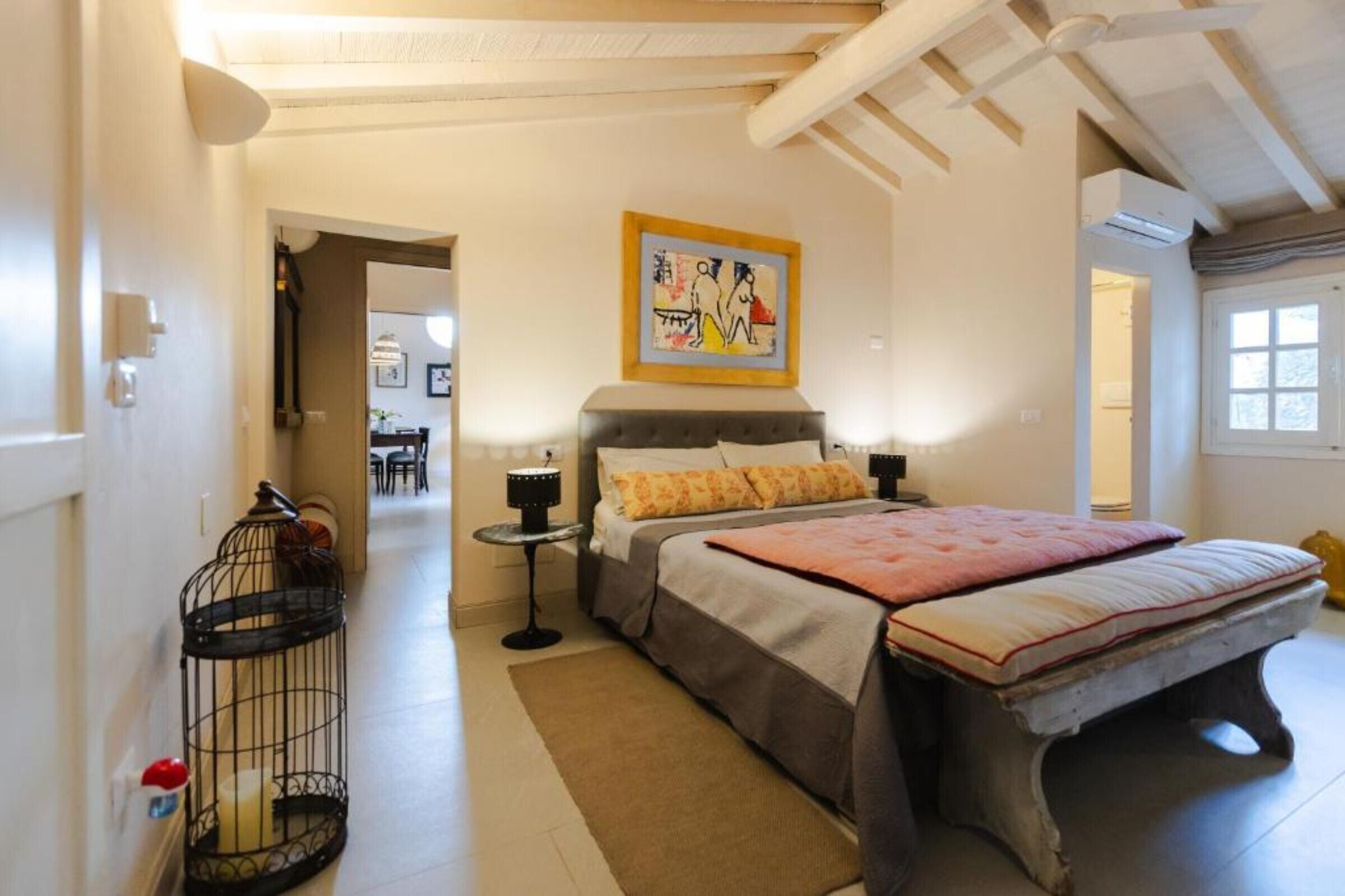 A bedroom with a bed, bedside tables, a nightstand, a painting, a window, and a door.