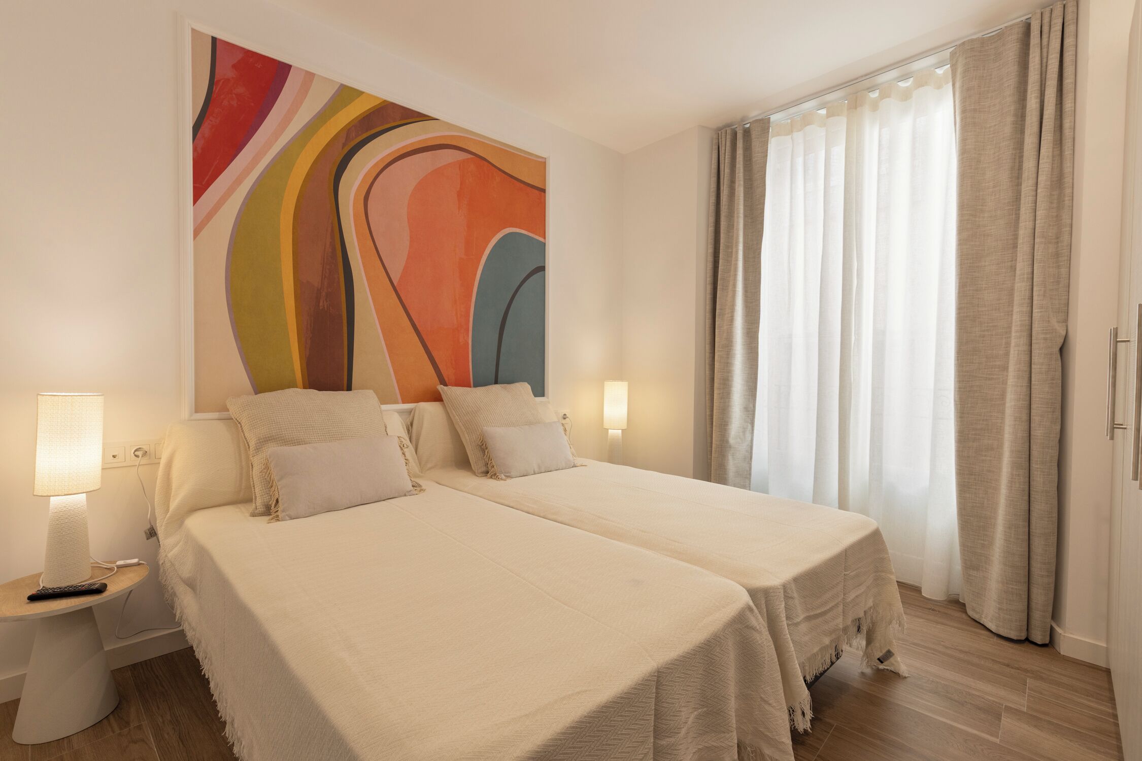 A hotel room with two beds, a large abstract painting on the wall, a nightstand with a lamp, and a window with curtains.