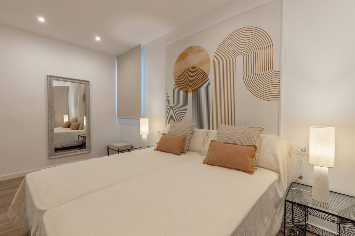 Un dormitorio moderno con una cama grande, una mesita de noche con lámpara, un espejo en la pared y arte abstracto en la pared.
