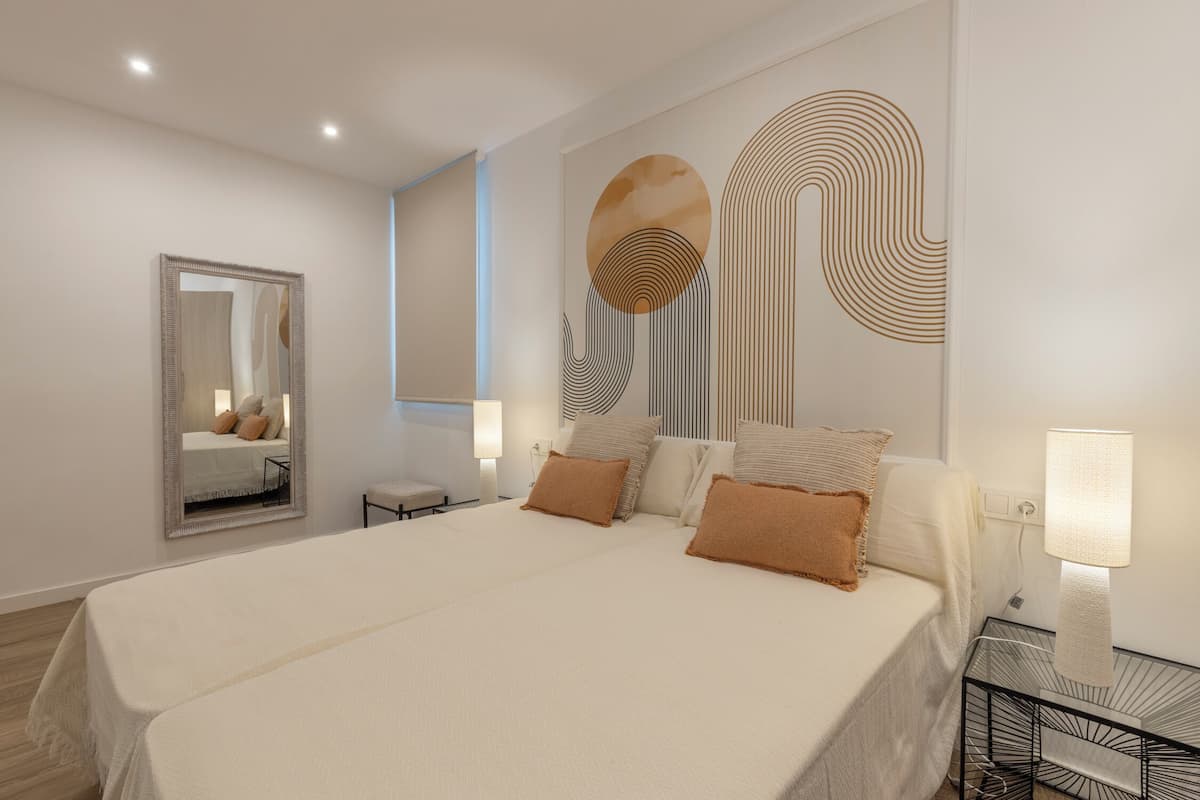 Une chambre moderne avec un grand lit, une table de chevet avec une lampe, un miroir fixé au mur et des œuvres d’art abstraites accrochées au mur.