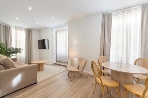 A modern living room with a sofa, a round dining table with chairs, a TV mounted on the wall, and large windows with curtains. - BIBO SUITES CASA NAWAL (Granada)