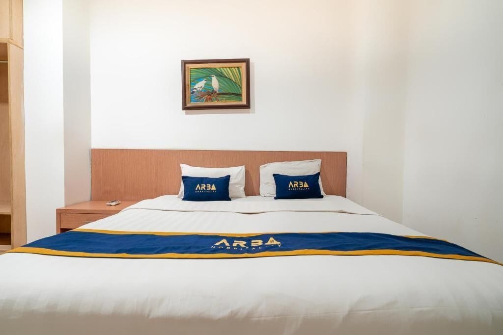 A hotel room with a bed, two pillows with the logo 'ARBA', a framed picture on the wall, and a bedside table with a phone.