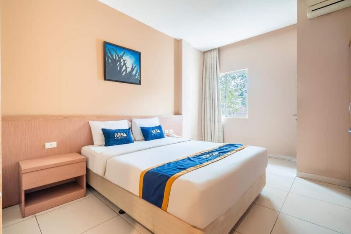 Ein Hotelzimmer mit einem Bett, einem Nachttisch, einem Wandbild, einem Fenster mit Vorhängen und einer Tür.